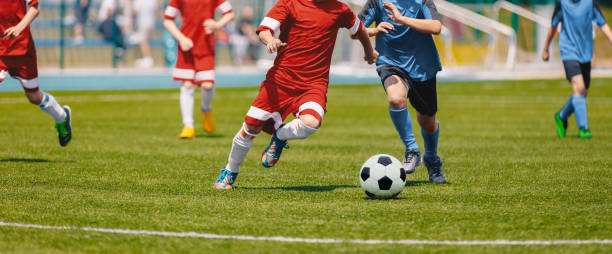 football soccer players running with ball. footballers kicking football match. young soccer players running after the ball. kids in soccer red and blue uniforms. soccer stadium in the background - soccer stadium soccer field sport imagens e fotografias de stock