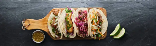 four mexican street tacos with fish barbacoa and carnitas shot in panoramic composition on top of serving wooden board