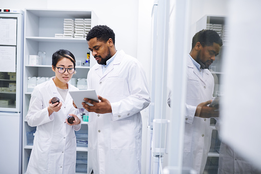 Content experienced multi-ethnic pharmaceutical workers in lab coats standing in storage room with cabinets and using tablet while comparing information about pills