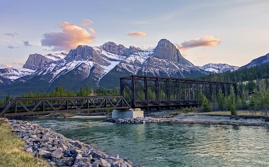 Historic Engine Bridge, a Steel Truss Pedestrian Footbridge, over Bow River in Canmore with Snowcapped Mountain Peaks Landscape on the Horizon