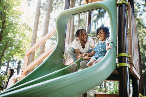 A dad assists his father going down a slide at a public city park in Tacoma, Washington.  They smile and laugh, having fun together in the warm weather.