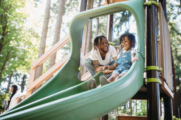 Father Helping His Child On Playground Slide A dad assists his father going down a slide at a public city park in Tacoma, Washington.  They smile and laugh, having fun together in the warm weather. tacoma stock pictures, royalty-free photos & images