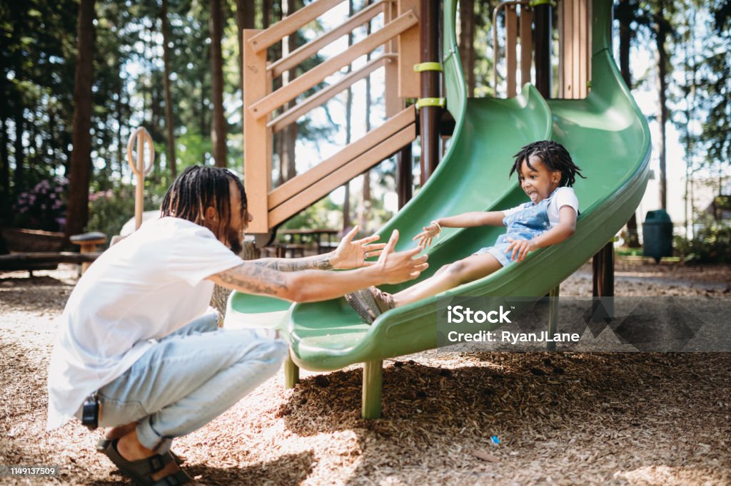 Father Helping His Child On Playground Slide A dad assists his father going down a slide at a public city park in Tacoma, Washington.  They smile and laugh, having fun together in the warm weather. Slide - Play Equipment Stock Photo