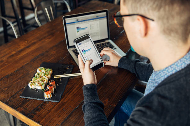 Millennial Checking Finances on Smart Phone A man of the Millennial Generation is looking at his financial statement while eating lunch at a local sushi restaurant. He is using his bank's app on his smart phone and laptop to balance his monthly budget and plan for the future. Image taken in Utah, USA. man looking at phone over shoulder stock pictures, royalty-free photos & images