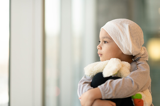 A beautiful little girl with cancer takes a break from treatment. She is sitting near a large bay of windows in the hospital's corridor. The girl is wearing a headscarf and is hugging a stuffed rabbit toy. She is looking out the window with a peaceful expression.