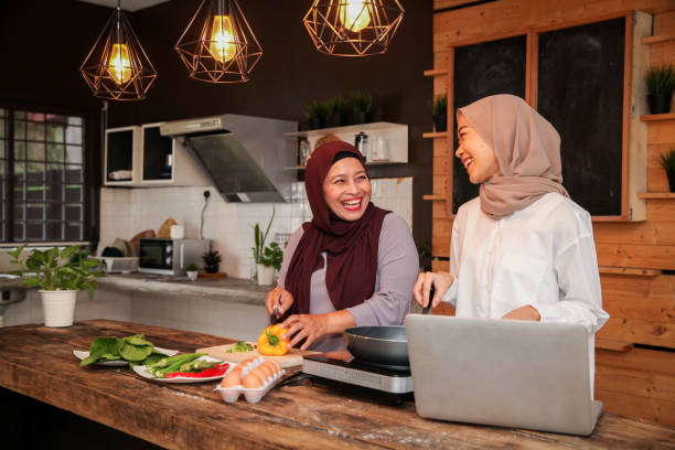Malay Muslim Mother and Daughter Refering to Laptop in the Kitchen Malay Muslim mother and daughter in hijab referring to laptop in the kitchen while preparing and cooking food for Hari Raya Aidilfitri/ Eid-Ul-Fitr hari raya family stock pictures, royalty-free photos & images