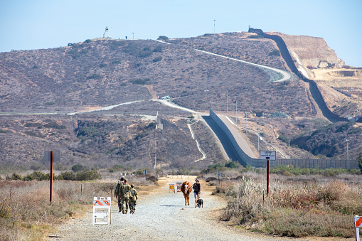 San Diego, California, USA - November 3, 2018 : US Armed Forces soldiers in green military camouflage walk near double-layered secured international border wall between the United States and Mexico
