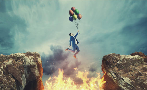 Photo of Businessman hold balloons and jumps over a fire between a gap of two mountain peaks.
