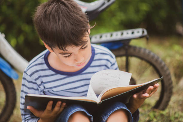 istock Little boy reading book sitting with bicycle in the park 1149186132