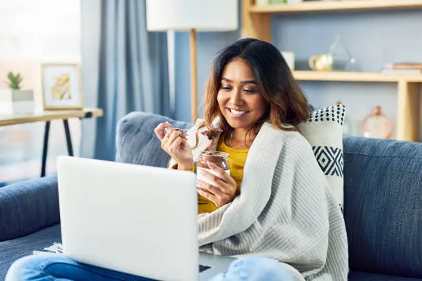 Shot of a woman looking at something on her laptop while eating chocolate from a jar