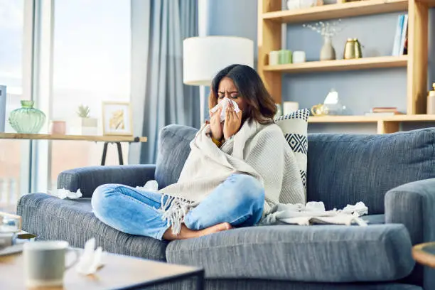 Shot of a young woman blowing her nose while sitting at home