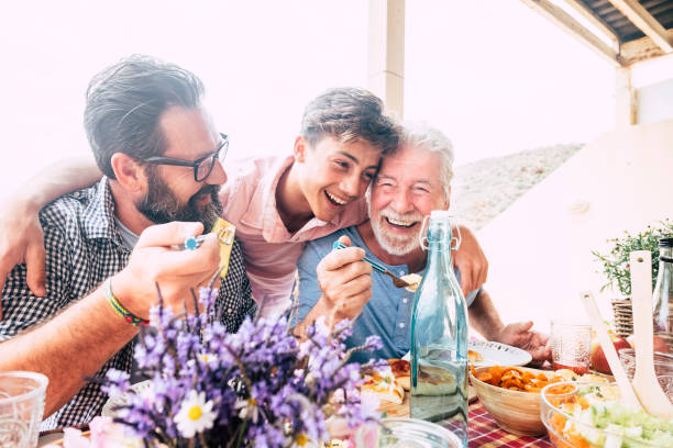 la familia de las personas felices se ríen y se divierten junto con tres generaciones diferentes: padre abuelo y joven hijo adolescente todos juntos comiendo en el almuerzo - bancal fotografías e imágenes de stock