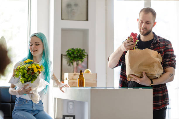 Woman with bunch of flowers and flatmate with groceries Anime-like young woman with bunch of flowers sitting at kitchen bar counter, flatmate taking radish out of grocery bag flatmate stock pictures, royalty-free photos & images