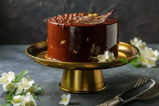 Chocolate mousse cake in mirror glaze on a bronze stand, on a dark textured background, selective focus.