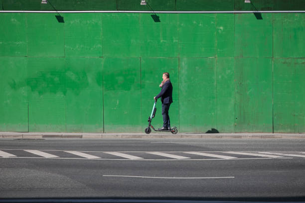 Copenhagen, Denmark – May 11, 2019:  Man on a scooter in front of a green fence. Scooters are a new and modern way of transport in Denmark and fences are a common sight in Copenhagen due to construction works
