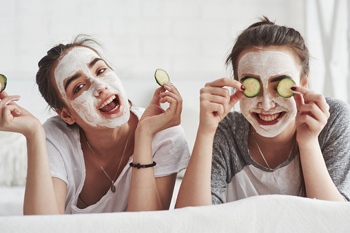 Nice day. Conception of skin care by using fresh cucumber rings and white mask on the face. Two female sisters have weekend at bedroom.