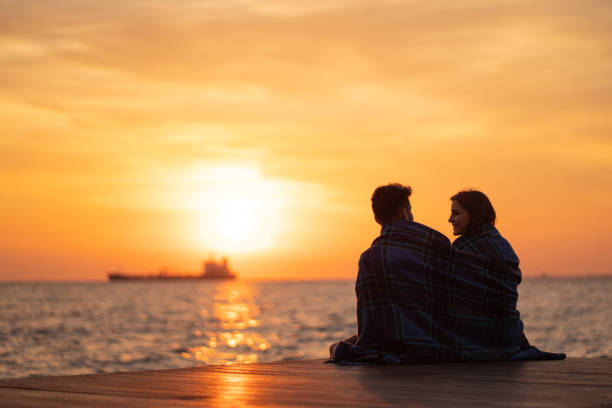 Young couple is sitting by the sea, wrapped in blanket, and looking amazing sunset.