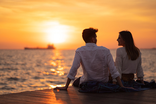 Young couple is sitting by the sea and looking amazing sunset.