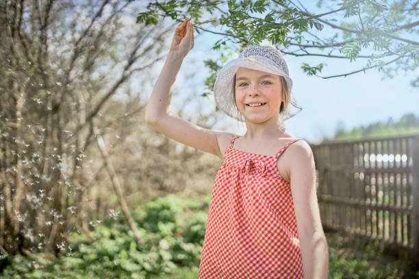 A beautiful elementary age girl dressed in a lace panama and bright summer clothes spends a weekend in a rural cottage. She is holding the dandelion, waving it playing with dandelion fluff. The girl is smiling and looking at the camera. Shooting on a back yard of a country cottage at a sunny spring day