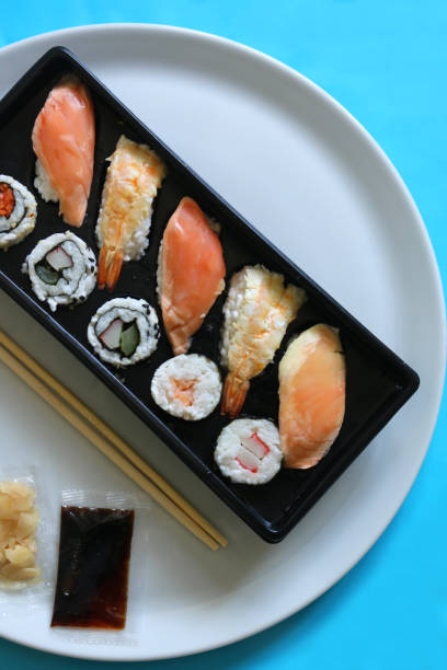 Stock photo of fresh sushi platter on white dinner plate, freshly prepared Japanese seafood sushi makizushi / maki ready to be eaten with soya sauce, wasabi sauce and bamboo chopsticks. imagen de restaurante japonés plato de sushi, mariscos recién preparados, tipos de sushi japón-makizushi, hosomaki/maki, futomaki, uramaki, temaki, nigiri, gunkanmaki, sashimi, salmón fresco, atún, colas de camarones, gambas, arroz pegajoso, envoltura
