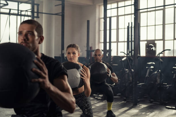 Fitness class using heavy weight balls Group of three people doing squats while holding heavy medecine balls. Young men and fitness woman doing bodyweight exercises using weight ball. People in a row squatting with weighted balls at cross training gym. people in a row photos stock pictures, royalty-free photos & images