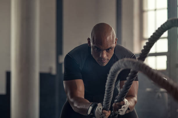 Athletic man using battle rope at gym Man in sportswear doing battle ropes functional training at cross training centre. Determined trainer making waves with ropes while exercising strength. Athlete working out with battle rope at industrial gym. effort stock pictures, royalty-free photos & images