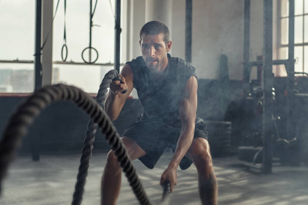 Young man exercising using battle rope Strong young man working out with battle ropes in a cross training gym. Muscular sportsman doing cross excursion with ropes in workout gym. Determined guy using battle rope while doing physical training. gym men africa muscular build stock pictures, royalty-free photos & images