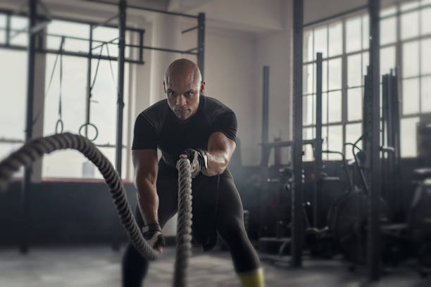 Mature strong man battling with rope Athlete working out with battle rope at gym. Bald african man training using battle ropes. Fit sportsman doing cross training exercise in an industrial dark gym. athlete stock pictures, royalty-free photos & images