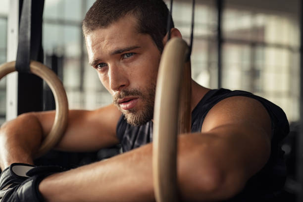 Closeup of young man resting after gymnastic ring exercise at cross training gym. Fitness guy thinking while leaning at dip rings and looking away. Determined man relaxing after workout training.
