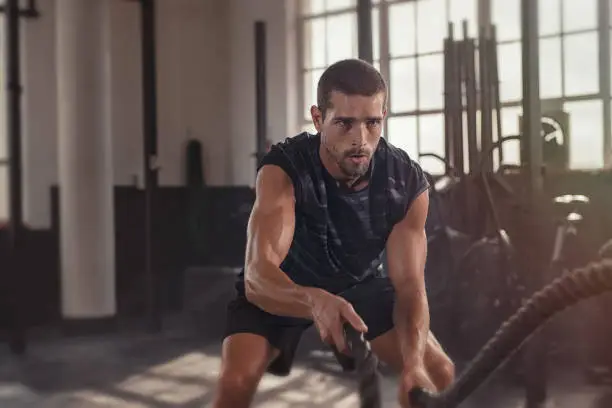 Athletic young man doing some cross training exercises with a rope. Determined fit guy doing battle ropes exercise at the cross training gym. Handsome man training with effort.