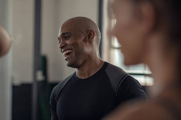 Mature fitness man laughing in gym Bald mature man enjoying a break while chatting at gym. Closeup of african american athlete laughing while in conversation with woman in fitness center. Carefree black trainer in sportswear resting after workout exercise with class. gym men africa muscular build stock pictures, royalty-free photos & images