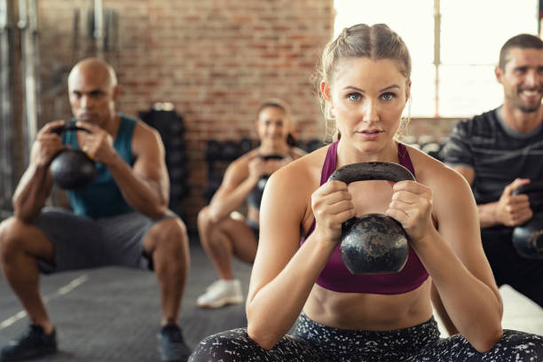 Fitness woman squatting with kettle bell Group of fit people holding kettle bell during squatting exercise at cross training gym. Fitness girl and men lifting kettlebell during strength training exercising. Group of young people doing squat with kettle bell. squatting position photos stock pictures, royalty-free photos & images