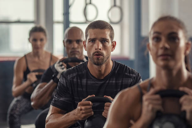 Fitness people exercising using kettle bell Portrait of fitness man lifting kettle bell with young women in fitness center. Active guy with group of people in gym doing weight lifting with kettlebell. Determined people in a row squatting in gym gym. people in a row photos stock pictures, royalty-free photos & images