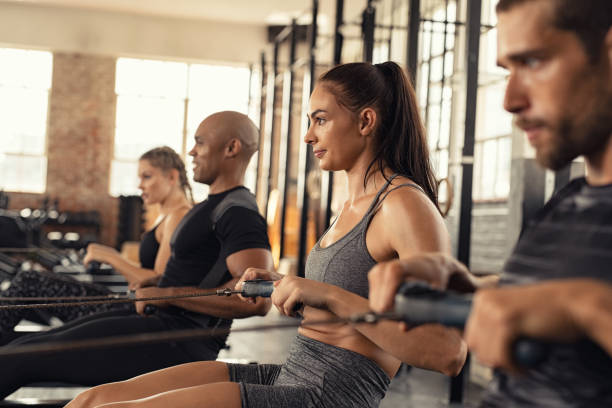 Fitness class training on rowing machine Group of people exercising in gym using rowing machine together. Side view of sportswoman doing exercise on rowing machine in cross training center. Muscular girl and sporty men workout on training simulator at gym gym. cardiovascular exercise stock pictures, royalty-free photos & images