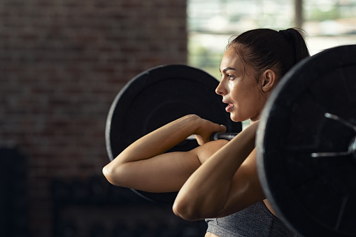 Fitness woman doing shoulder press exercise with a weight bar at cross training gym. Muscular woman in gym doing heavy weight exercises. Concentrated athlete doing barbell lifting at health club with copy space.