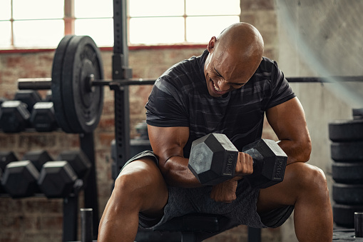 Muscular guy in sportswear lifting dumbbell while sitting on bench at cross training gym. Mature african american athlete using dumbbell during a workout. Strong man under physical exertion pumping up bicep muscule with heavy weight.