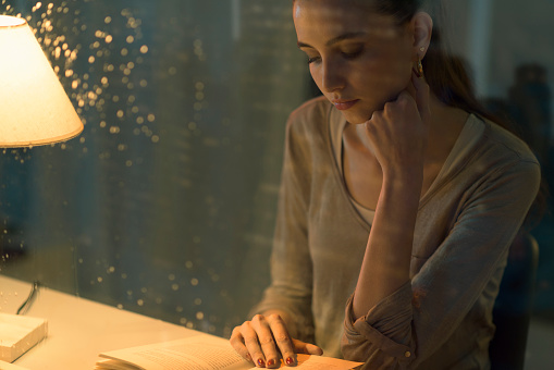 Young woman reading a book at night, she is sitting at desk, glass with raindrops in the foreground