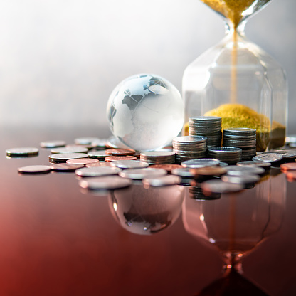 Global business investment growth. Wealth concept. Gold sand running through the shape of modern hourglass with world globe crystal glass and US dollar coins on red reflective table.