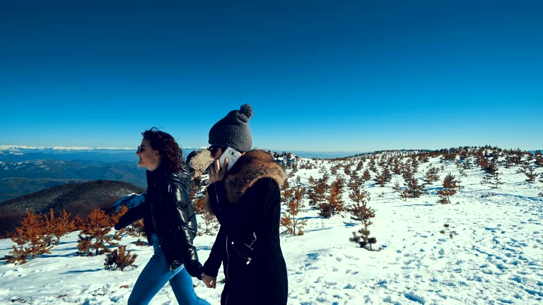 Two female teens walking on snow holding hands and talking on smartphone smiling