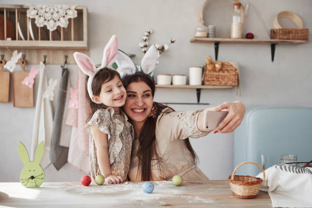 istock Using white colored smartphone. Mother and daughter in bunny ears at easter time have some fun in the kitchen at daytime 1149277283