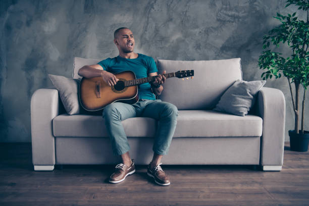 vollständige körpergröße foto erstaunlich er ihn seine dunkle haut macho halten arme gitarre gesang solisten spielen allein rock legenden hits tragen blaue t-shirt hosen sitzen bequeme divan raum bürohaus in innenräumen - skin singer stock-fotos und bilder