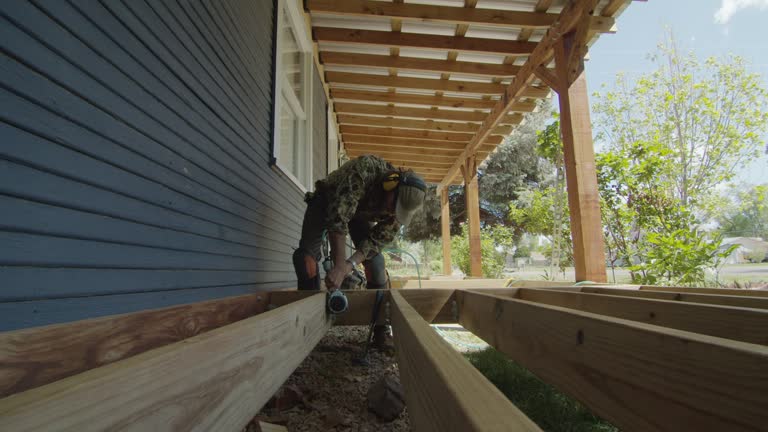 A Caucasian Handyman in His Forties Wearing a Hat, Hearing Protection, and Kneepads Uses a Pneumatic Nail Gun to Secure Metal Corner Brackets on Wooden Boards while Building a Deck in a Residential Neighborhood Outdoors on a Sunny Day