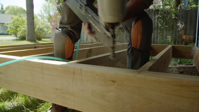 A Caucasian Handyman in His Forties Wearing a Hat, Hearing Protection, and Kneepads Uses a Pneumatic Nail Gun to Position a Wooden Board and then Uses It to Secure the Board into Place Then Uses a Hammer While Building a Deck in a Residential Neighborhood