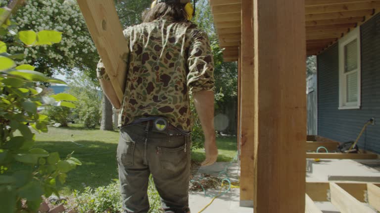 A Caucasian Handyman in His Forties Wearing a Hat, Hearing Protection, and Kneepads Carries a Wooden Board while Building a Deck in a Residential Neighborhood Outdoors on a Sunny Day