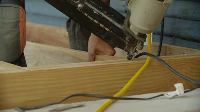 A Caucasian Handyman Wearing Kneepads Uses a Pneumatic Nail Gun to Attach Wooden Boards While Building a Deck in a Residential Neighborhood Outdoors on a Sunny Day