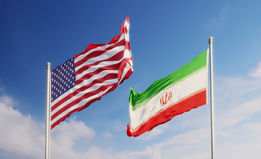 American and Iranian flags waving at opposite directions over blue sky. Horizontal composition with copy space.