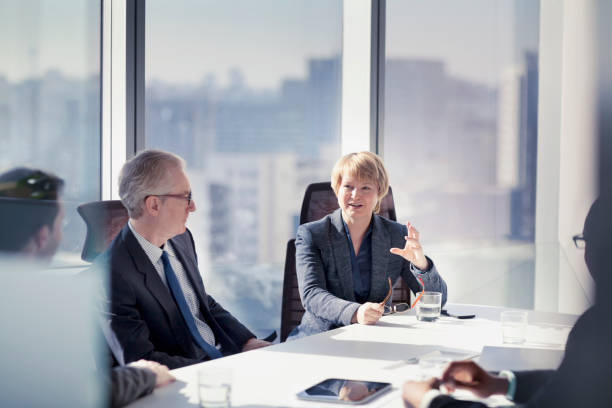 Businesswoman talking with colleagues in meeting Mature adult businesswoman leader boss CEO executive with group of business people sitting together in conference meeting room in contemporary modern office bright sunny daylight talking discussing planning organizing strategy well dressed suit and tie teamwork cooperation diversity multi-ethnic problems solutions unity cityscape downtown urban formal businesswear stock pictures, royalty-free photos & images