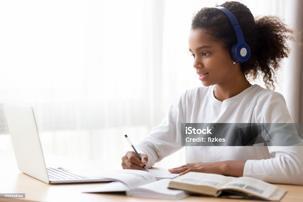 African American teen girl wearing headphones learning language online African American teen girl wearing headphones learning language online, using laptop, looking at screen, doing school tasks at home, writing notes, listening to lecture or music, distance education Child Stock Photo