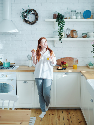 Beautiful authentic young woman with red hair enjoying eating pastry and drinking orange juice for breakfast and day dreaming in sunlight leaning on counter in kitchen