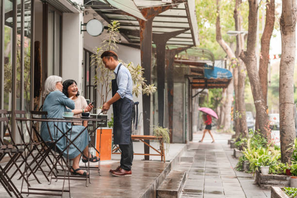Senior women using contactless payment at a cafe Two senior women are using their phone to order and pay for their coffee at a cafe. The waiter is using a digital tablet. rainy cafe stock pictures, royalty-free photos & images